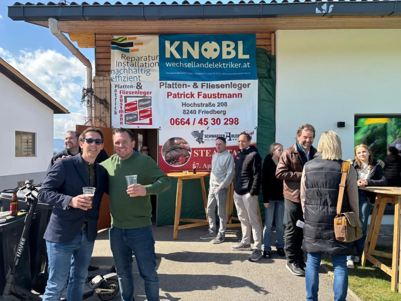 Eine Gruppe von Männern und Frauen steht vor einem Gebäude mit einem Schild für Platten- & Fliesenleger Patrick Faustmann. Das Schild enthält eine Telefonnummer und Adresse. Die Männer lächeln und halten Getränke in den Händen.