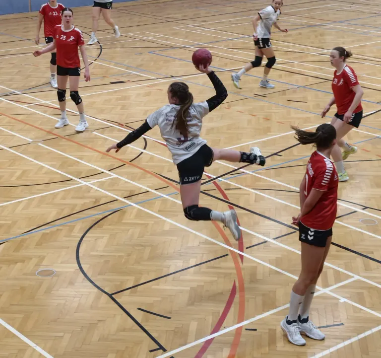 Eine Gruppe von Sportlerinnen spielt Volleyball auf einem Hallenplatz. Eine Spielerin in der Vordergrund springt, um den Ball zu schlagen. Andere Spielerinnen sind in Position, um zu verteidigen oder anzugreifen.
