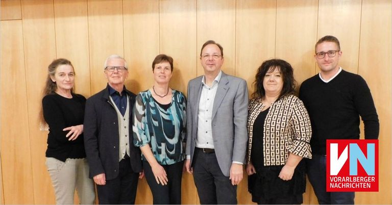Sechs Personen stehen gemeinsam und posieren für ein Foto vor einer Holzwand. Sie sind in formeller Kleidung gekleidet.