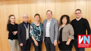 Sechs Personen stehen gemeinsam und posieren für ein Foto vor einer Holzwand. Sie sind in formeller Kleidung gekleidet.