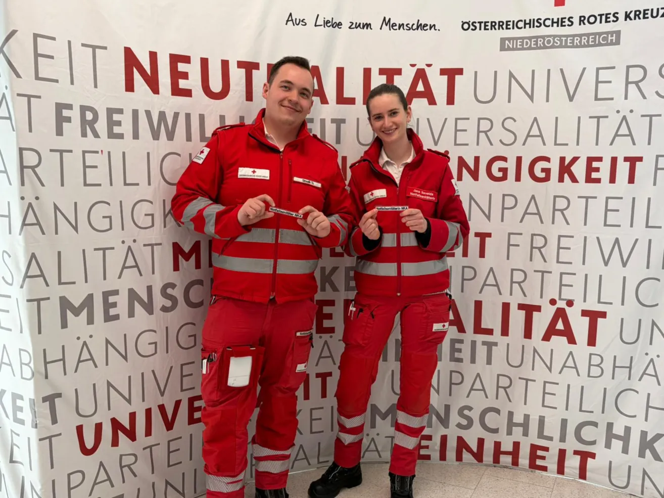 Zwei Personen in roten Uniformen, die Ausweise halten, stehen vor einem Hintergrund mit dem Wort 'NEUTRALITAT' in großen Buchstaben. Der Mann auf der linken Seite lächelt, während die Frau auf der rechten Seite ihren Ausweis mit beiden Händen hält.