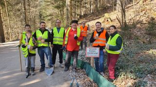 Eine Gruppe von Menschen in Sicherheitswesten mit Tassen und Schaufeln steht auf einem Kiesweg. Ein Schild warnt vor einem Froschübergang.
