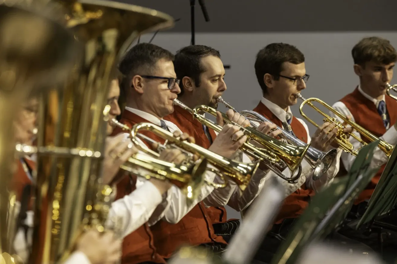 Vier Männer in roten Westen spielen Blasinstrumente in einem Raum. Drei von ihnen spielen Trompeten, während der andere ein Tuba spielt.