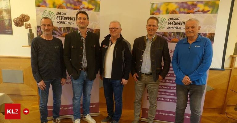 Fünf Männer stehen vor Bannern, die Gesundheit und Zukunft fördern. Sie tragen alle Freizeitkleidung und scheinen zu lächeln. Die Banner haben einen lila Hintergrund mit weißem Text.