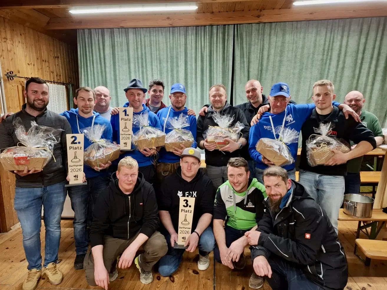 Eine Gruppe von Männern in blauen und schwarzen Jacken, einer mit einem blauen Hut, posiert für ein Foto. Sie halten verpackte Geschenke und Trophäen. Dahinter befindet sich ein grüner Vorhang.