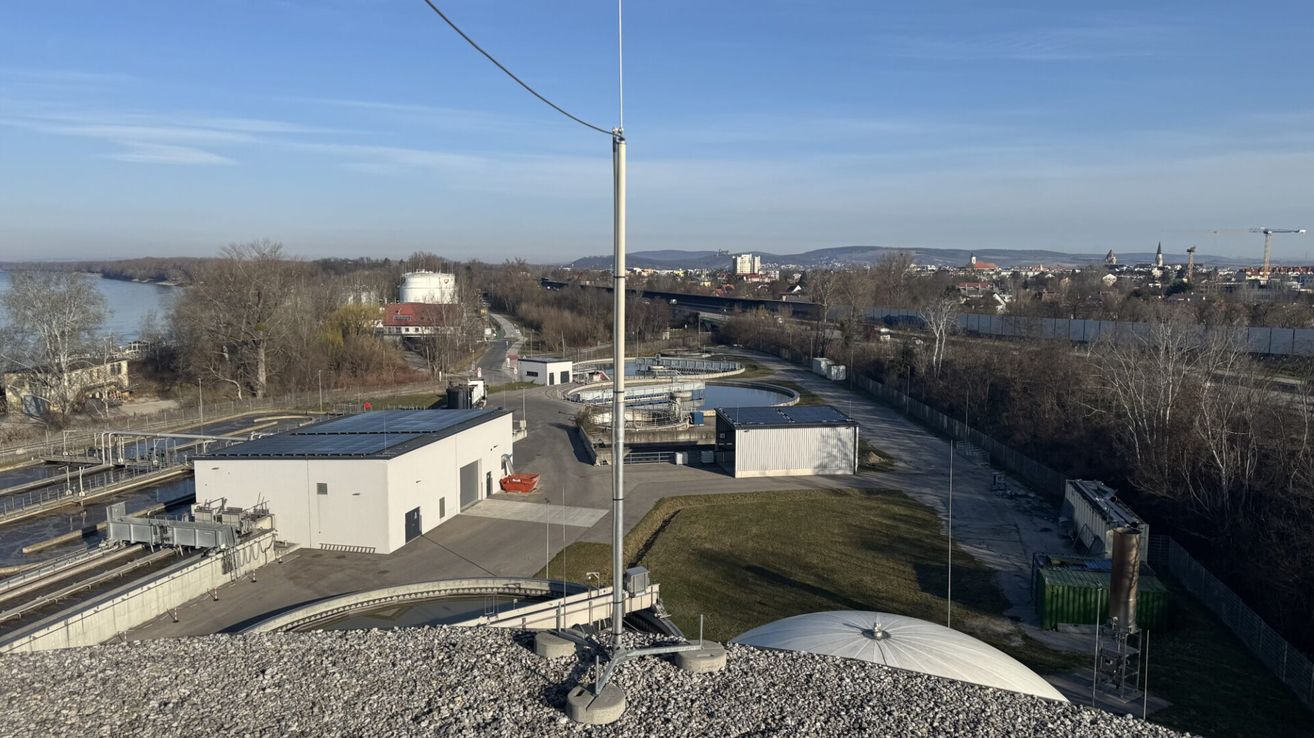 Luftaufnahme einer Kläranlage mit Gebäuden, Solarpanelen und einem großen Tank. Die Anlage ist von einer Kiesstraße umgeben und befindet sich in einem ländlichen Gebiet.