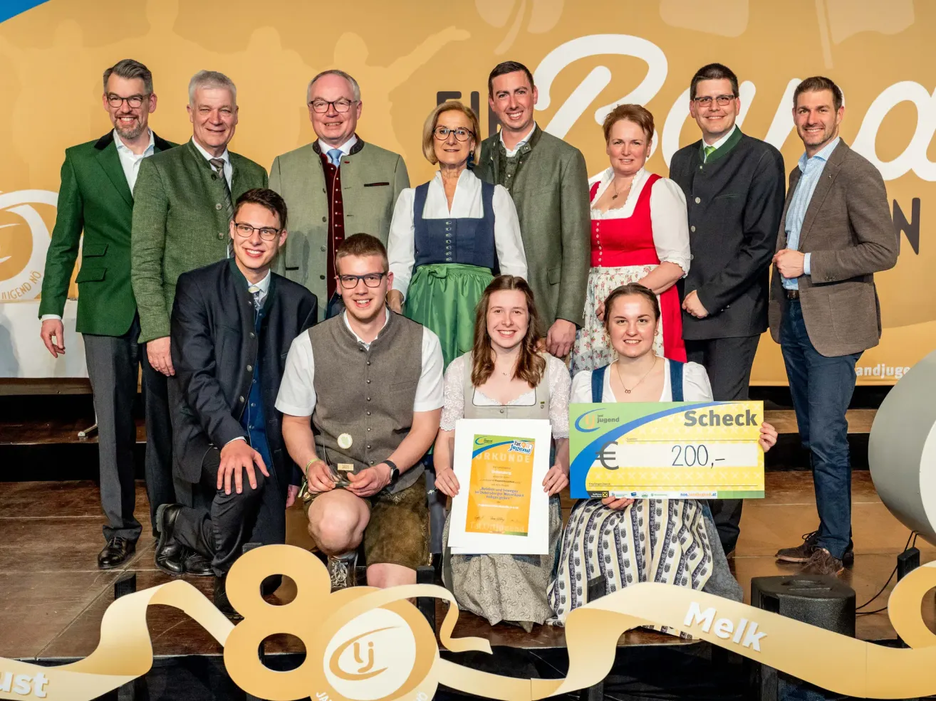 Eine Gruppe von Menschen steht auf einer Bühne, einige halten Auszeichnungen. Eine junge Frau hält einen großen Scheck, und andere halten Zertifikate. Sie tragen traditionelle Kleidung. Ein gelber Banner mit weißen Buchstaben ist hinter ihnen.