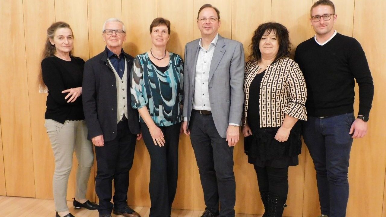 Eine Gruppe von fünf Personen steht vor einer Holzwand und lächelt für ein Foto. Sie sind formell gekleidet, mit zwei Frauen und drei Männern.