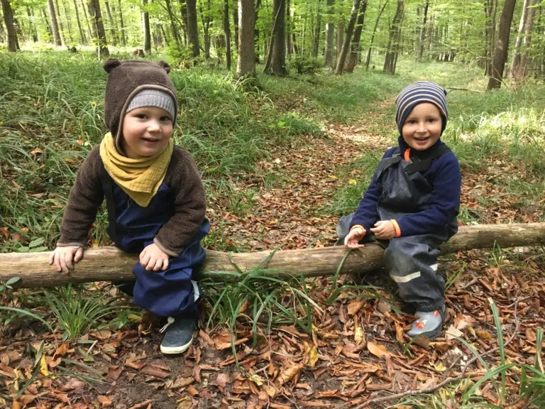 Zwei Jungen sitzen lächelnd auf einem Baumstamm im Wald, tragen warme Kleidung und Mützen. Der Boden ist mit Blättern und Gras bedeckt.