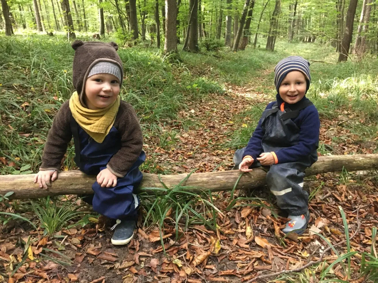 Zwei Jungen sitzen lächelnd auf einem Baumstamm im Wald, tragen warme Kleidung und Mützen. Der Boden ist mit Blättern und Gras bedeckt.