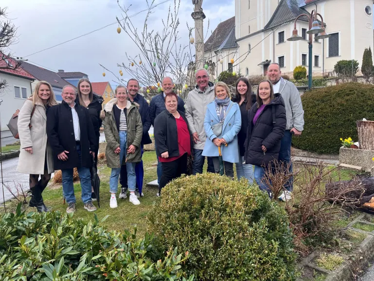Eine Gruppe Erwachsener posiert für ein Foto in einem Garten mit einem Gebäude im Hintergrund und einem bewölkten Himmel.