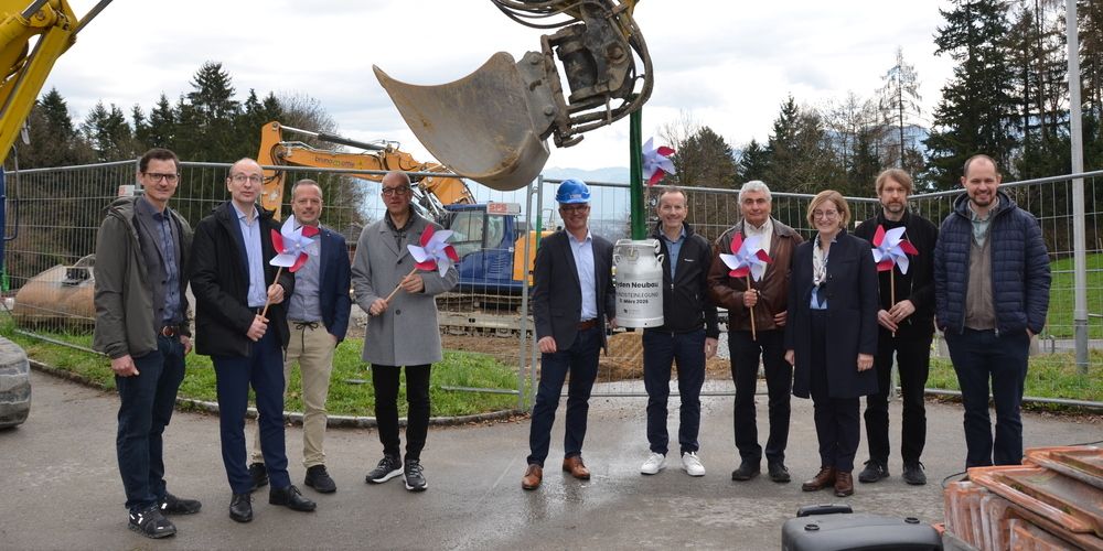 Mehrere Personen stehen in einer Gruppe vor einer Baustelle, halten Windräder und lächeln für die Kamera. Dahinter steht ein Baufahrzeug.