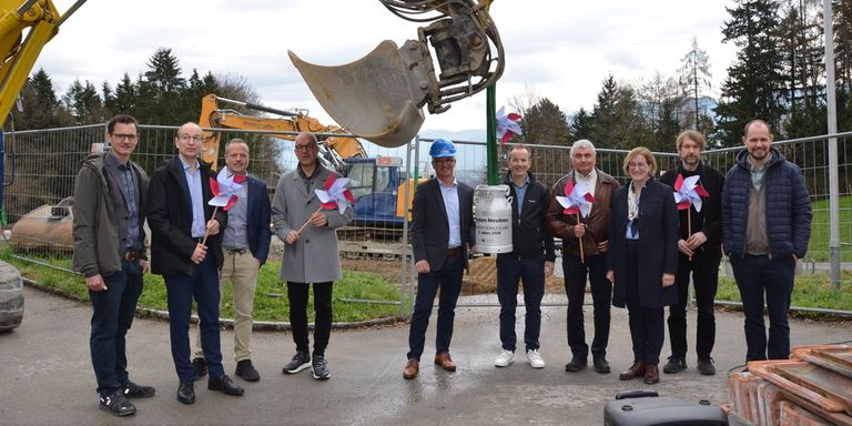 Mehrere Personen stehen in einer Gruppe vor einer Baustelle, halten Windräder und lächeln für die Kamera. Dahinter steht ein Baufahrzeug.