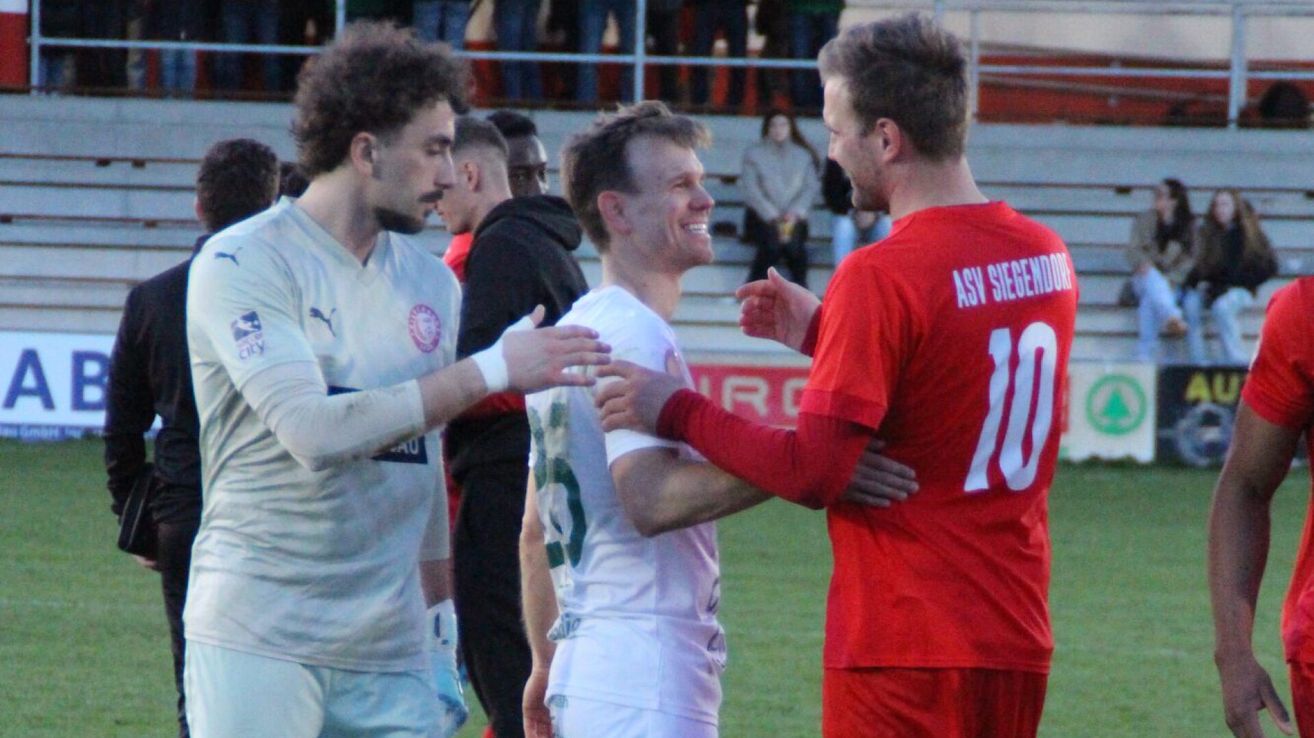 Drei Fußballspieler auf einem Feld, zwei in weißen Trikots und einer in Rot. Sie schütteln sich die Hände und lächeln, mit Zuschauern im Hintergrund.