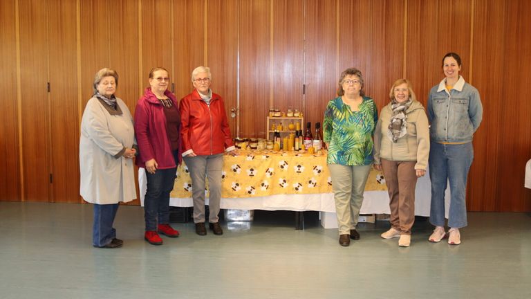 Fünf Frauen stehen hinter einem Tisch mit Gläsern und Flaschen. Der Tisch hat einen gelben Stoff mit Bienendesigns. Sie sind lässig gekleidet und lächeln. Hinter ihnen befindet sich eine Holzwand.