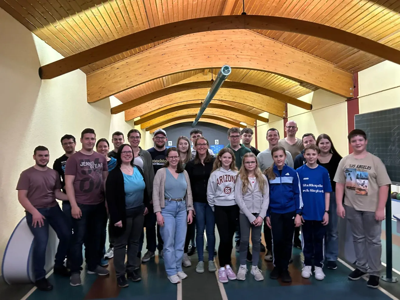 Eine Gruppe von Menschen in einem Flur, lächelnd für ein Foto. Sie tragen Freizeitkleidung und einige tragen Brillen. Die Decke ist aus Holz und in der Mitte befindet sich eine Rohrleitung. Die Wände sind weiß gestrichen.