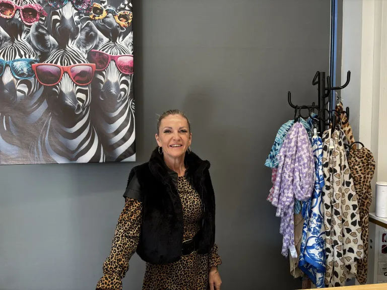 Eine Frau steht vor einer grauen Wand, die mit einem großen Zebra-Aufdruck verziert ist, und lächelt. Sie trägt ein Leopardenmusterkleid und eine schwarze Pelzweste. In der Nähe hängen Kleider an einem Metallbügel.