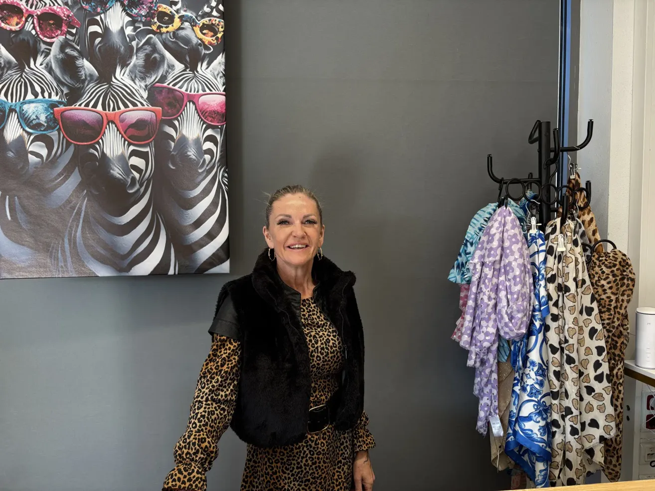 Eine Frau steht vor einer grauen Wand, die mit einem großen Zebra-Aufdruck verziert ist, und lächelt. Sie trägt ein Leopardenmusterkleid und eine schwarze Pelzweste. In der Nähe hängen Kleider an einem Metallbügel.