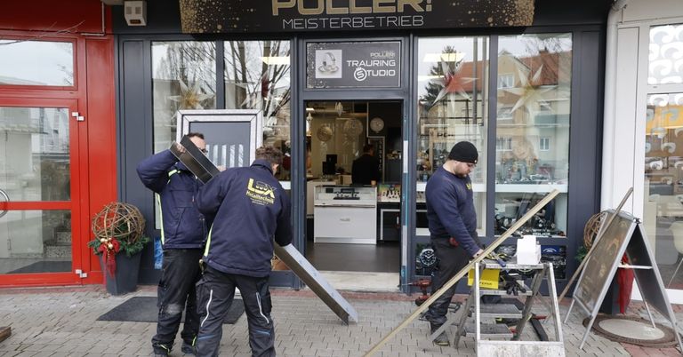 Drei Männer in blauen Jacken arbeiten vor einem Geschäft. Sie halten eine Metallplatte. Das Geschäft hat eine Glastür mit einem Schild, das 'PULLERI TRAURING STUDIO' besagt.