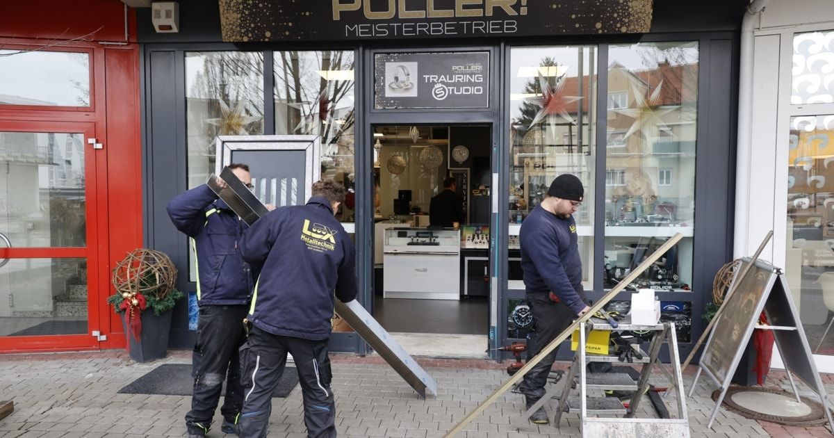Drei Männer in blauen Jacken arbeiten vor einem Geschäft. Sie halten eine Metallplatte. Das Geschäft hat eine Glastür mit einem Schild, das 'PULLERI TRAURING STUDIO' besagt.