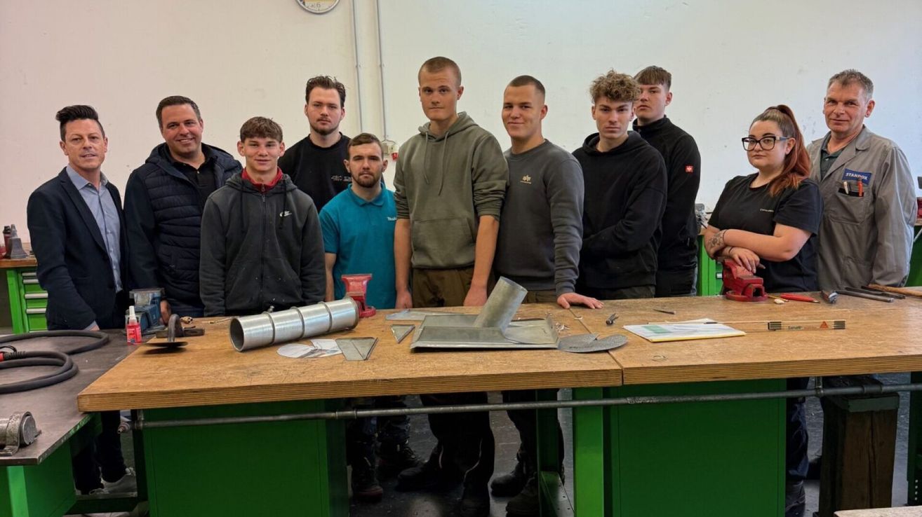 Eine Gruppe junger Männer in Freizeitkleidung steht in einer Werkstatt und posiert für ein Foto. Sie stehen um einen Tisch herum, auf dem Metallgegenstände und Werkzeuge liegen. Hinter ihnen befindet sich eine weiße Wand mit einer Uhr und einer Steckdose.