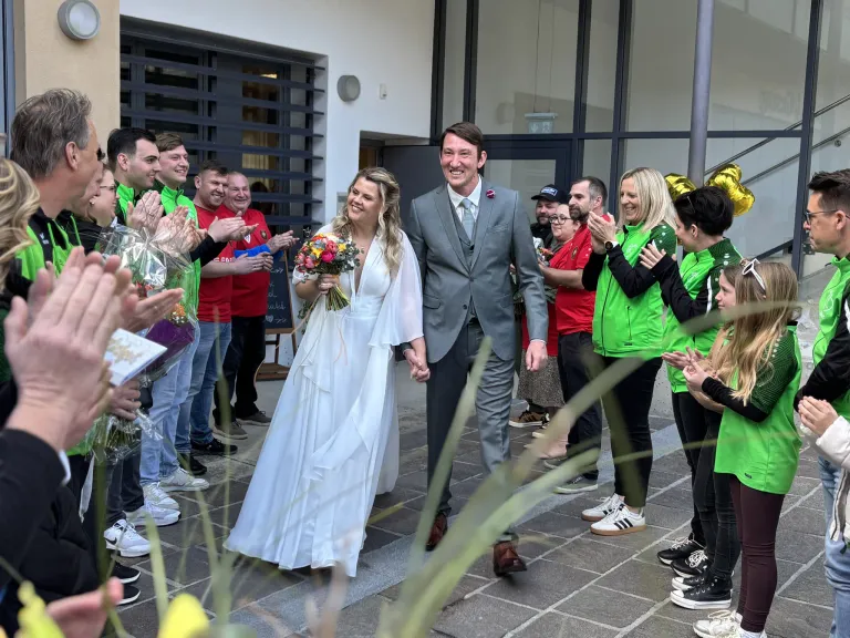 Ein frisch verheiratetes Paar geht lächelnd und Händchen haltend einen Pfad entlang, umgeben von einer Menge jubelnder und applaudierender Menschen.