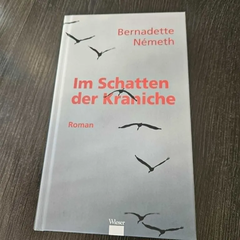 Ein Buch mit dem Titel 'Im Schatten der Kraniche' von Bernadette Nemeth. Das Cover zeigt Vögel, die über einem grauen Hintergrund fliegen. Das Buch liegt auf einer Holzoberfläche.