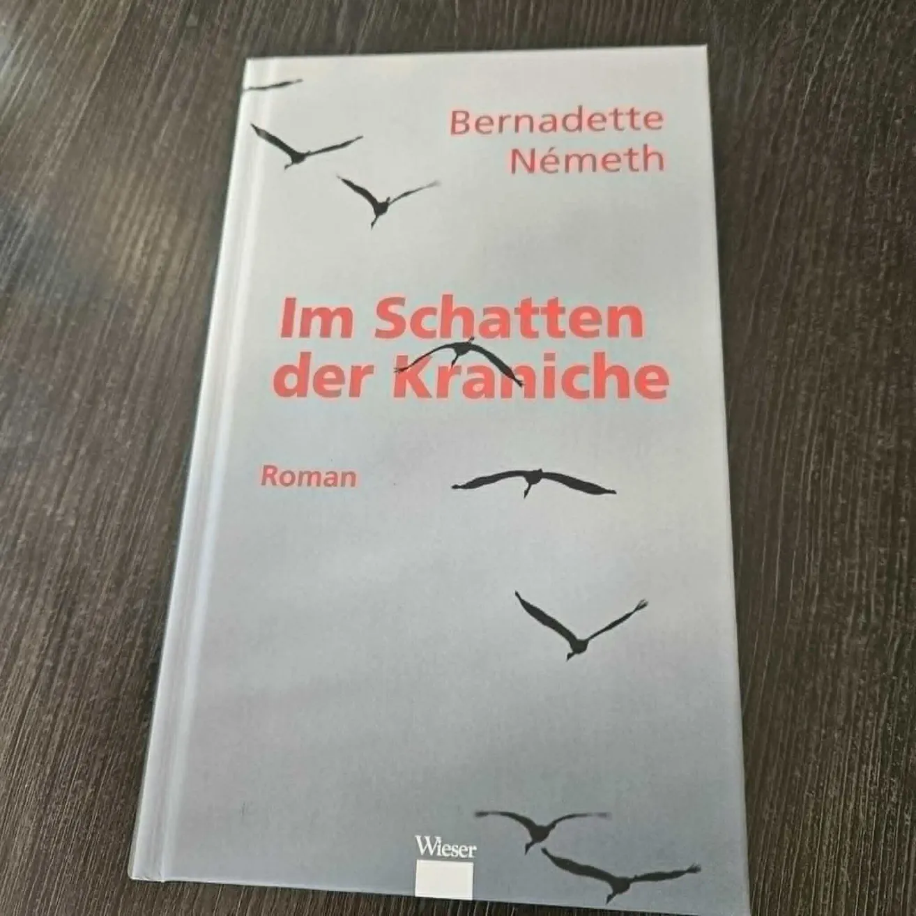 Ein Buch mit dem Titel 'Im Schatten der Kraniche' von Bernadette Nemeth. Das Cover zeigt Vögel, die über einem grauen Hintergrund fliegen. Das Buch liegt auf einer Holzoberfläche.