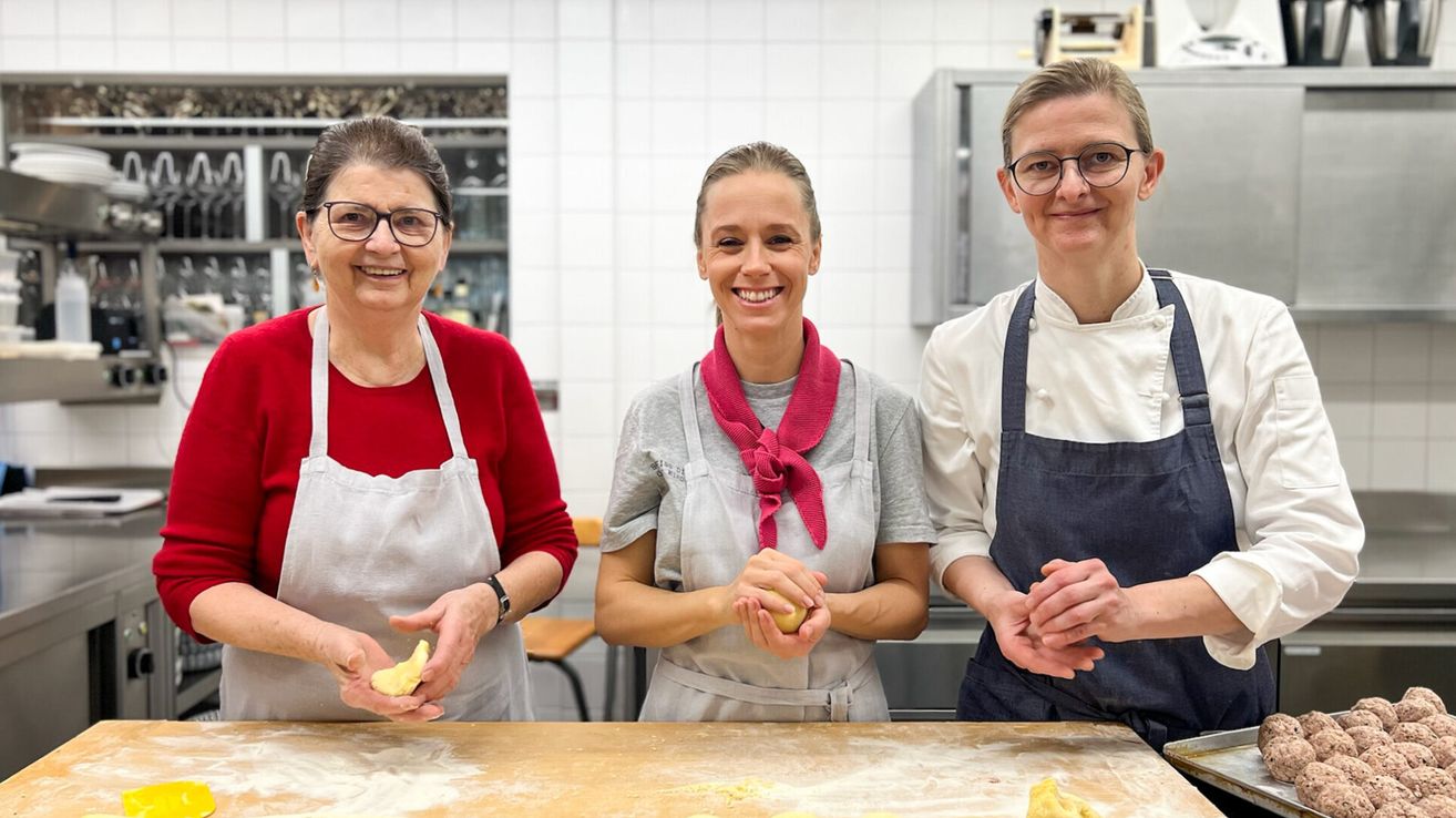 Drei Frauen in Schürzen stehen in einer Küche. Sie halten Teig und lächeln. Ein Tresen und Regale mit Gegenständen befinden sich hinter ihnen.