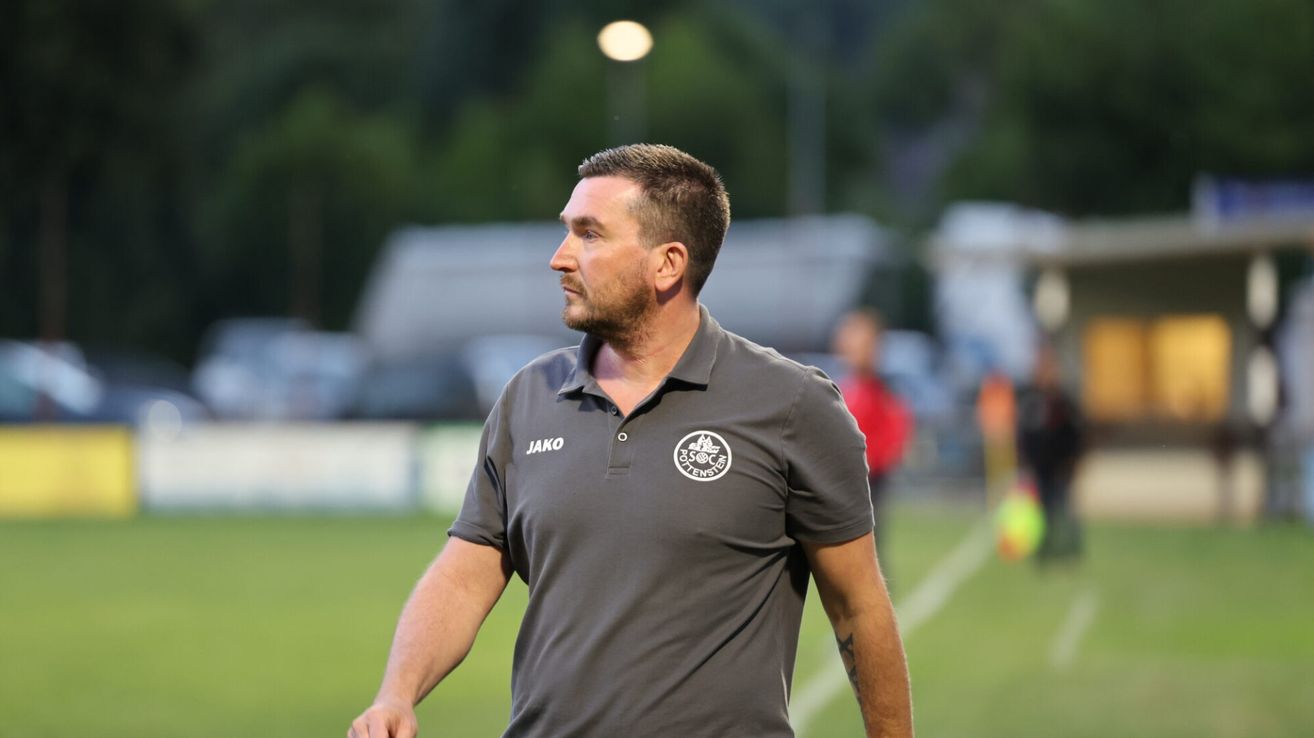 Ein Mann in einem grauen Poloshirt mit Logo steht auf einem Fußballfeld.