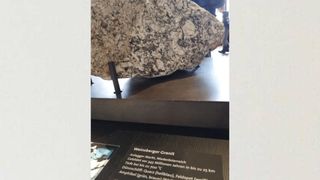 Ein großer Stein mit der Aufschrift 'Weinsberg Granit' wird in einem Museum ausgestellt. Der Stein stammt aus dem Ardaigger-Markt in Niederösterreich. Er entstand vor 345 Millionen Jahren und ist bis zu 700 Millionen Jahre alt. Er ist 25 km dick und enthält Amphibol (grün, braun), Quarz (hellblau) und Feldspat (weiß).