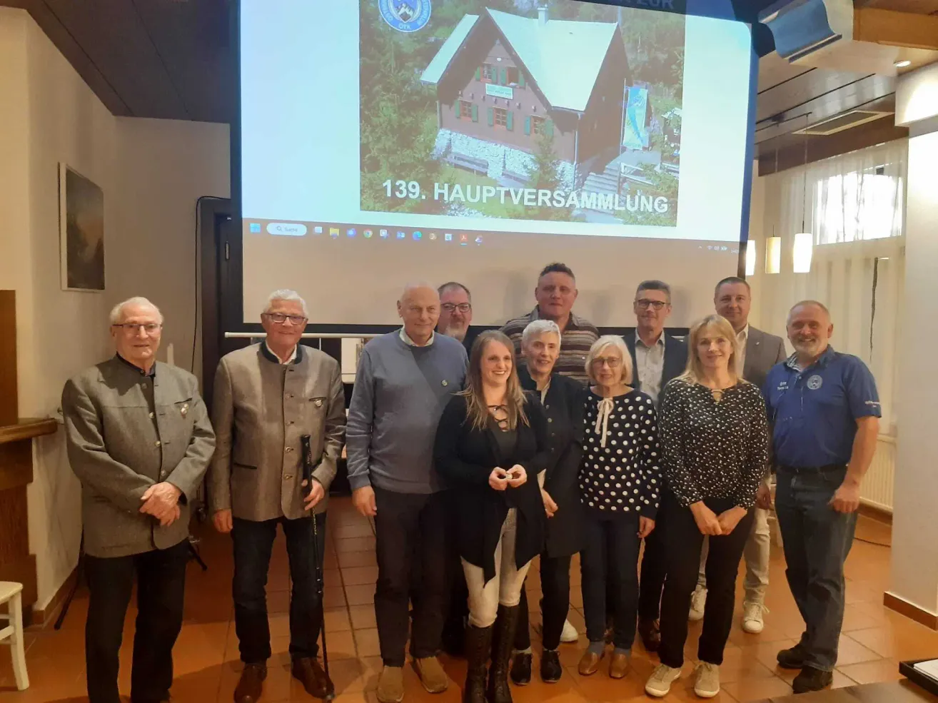 Eine Gruppe von Menschen posiert für ein Foto vor einer Projektionsleinwand, auf der ein Haus und die Worte 139 Hauptversammlung zu sehen sind.