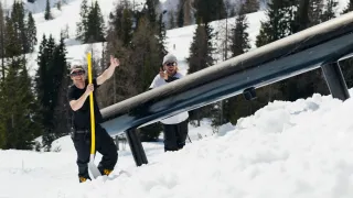 Zwei Männer in Skiausrüstung, einer mit einem gelben Stock, posieren für ein Foto in der Nähe einer Schneeröhre an einem verschneiten Hang mit Bäumen im Hintergrund.