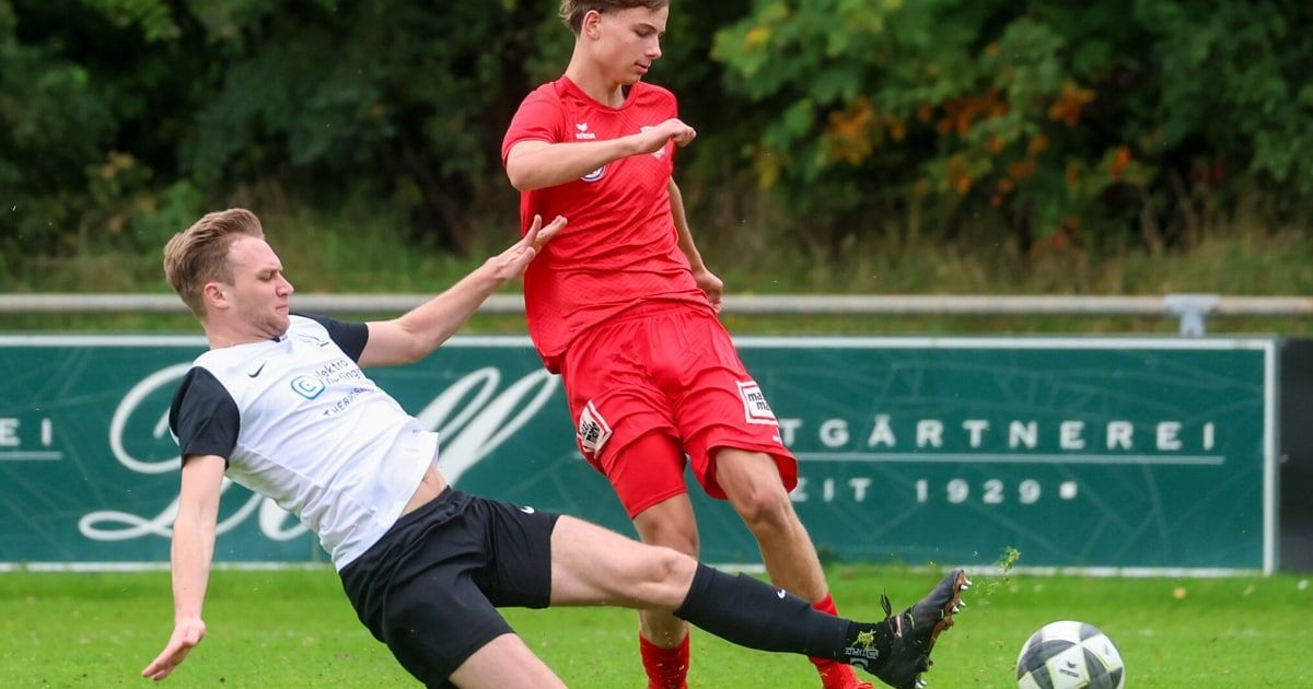 Zwei männliche Fußballspieler sind auf einem Feld in Aktion. Einer ist in Rot und versucht, einen Tritt des anderen in Weiß zu blockieren. Ein Fußball liegt in der Nähe.