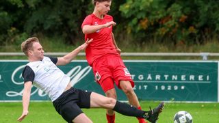 Zwei männliche Fußballspieler sind auf einem Feld in Aktion. Einer ist in Rot und versucht, einen Tritt des anderen in Weiß zu blockieren. Ein Fußball liegt in der Nähe.