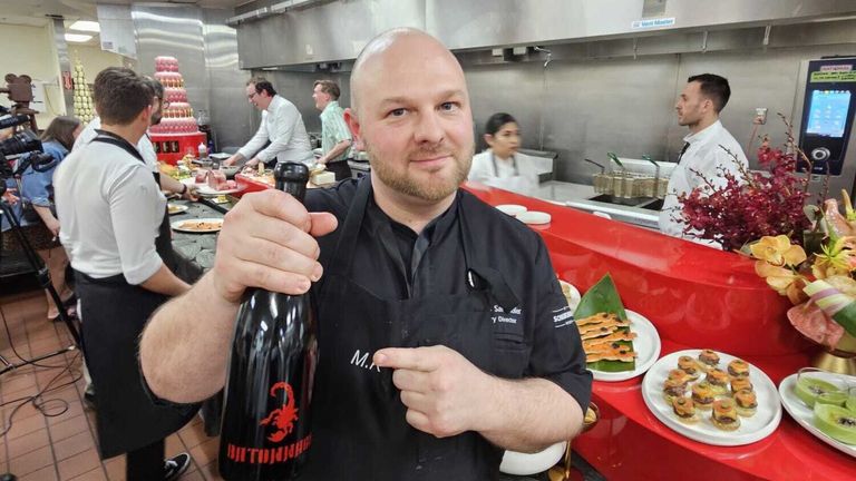 Ein Koch hält eine Flasche Scorpion-Wein in einer Restaurantküche. Andere Köche arbeiten hinter ihm. Teller mit Essen stehen auf der Theke.