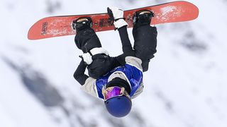 Ein Snowboarder befindet sich in der Luft und führt einen Trick mit einem roten Snowboard aus. Der Fahrer trägt einen blauen Helm, eine Schutzbrille und schwarze Hosen.