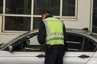 Ein Polizeibeamter in einer reflektierenden Weste steht neben einem silbernen Auto, möglicherweise zur Überprüfung oder Routinekontrolle.