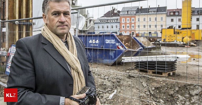 Ein Mann in Anzug und Schal hält eine Kamera vor einer Baustelle mit einem blauen Container und einem Gebäude hinter einem Zaun.