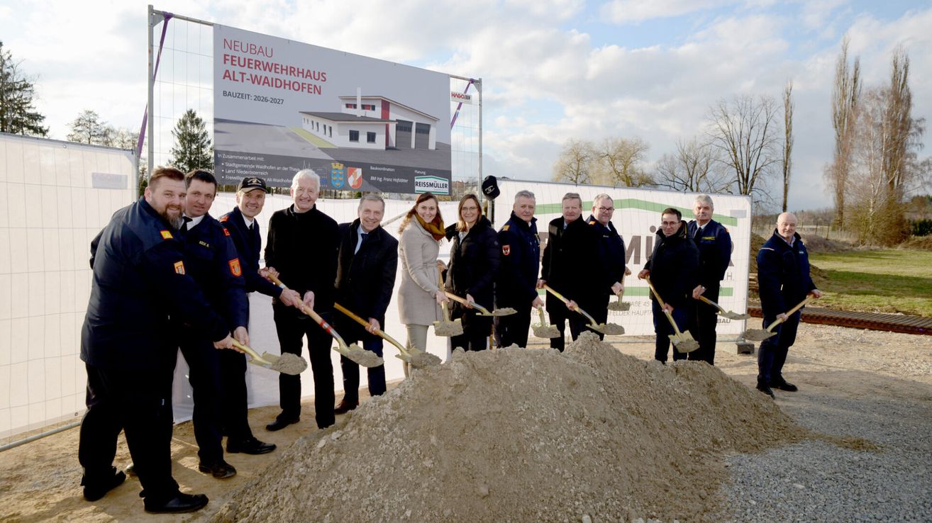 Eine Gruppe von Menschen in Uniform mit Schaufeln, die vor einem Haufen Erde stehen. Dahinter befindet sich ein großes Banner mit dem Text 'Neubau Feuerwehr Alt-Waidhofen'.
