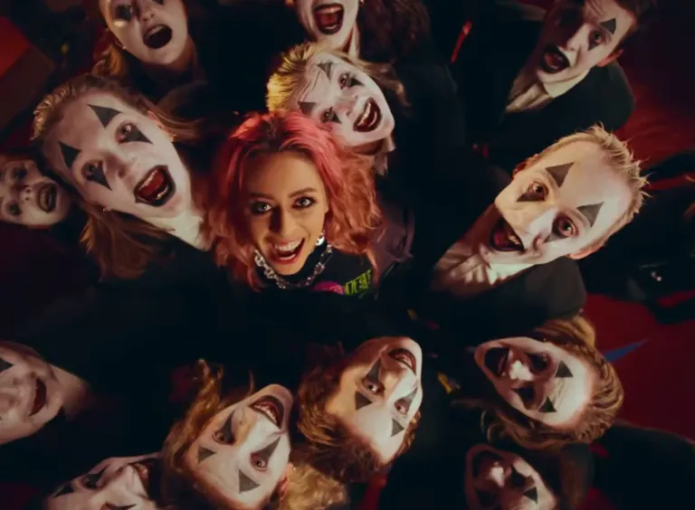 Eine Gruppe von Menschen mit Clown-Make-up, jeder mit einzigartigen Gesichtsausdrücken, umgibt eine Frau mit rosa Haaren. Der Hintergrund ist dunkel.