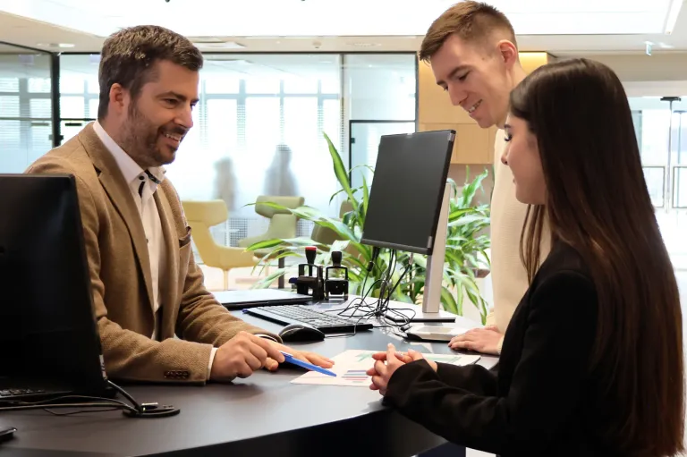 Drei Personen befinden sich in einer Büroumgebung. Ein Mann und eine Frau sitzen und unterhalten sich, während ein Mann hinter ihnen steht, lächelt und auf den Monitor schaut.
