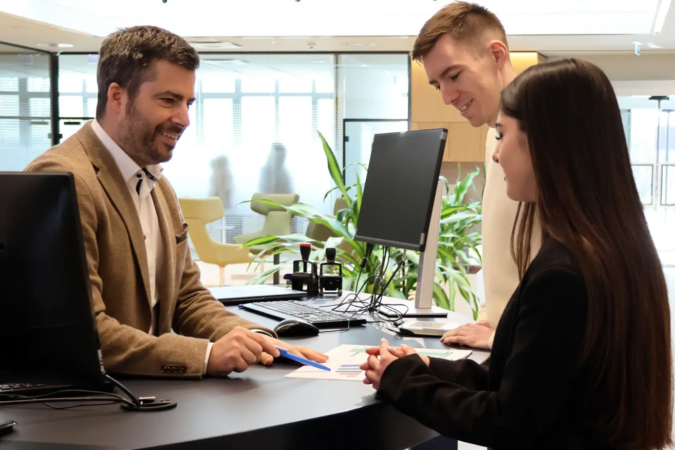 Drei Personen befinden sich in einer Büroumgebung. Ein Mann und eine Frau sitzen und unterhalten sich, während ein Mann hinter ihnen steht, lächelt und auf den Monitor schaut.
