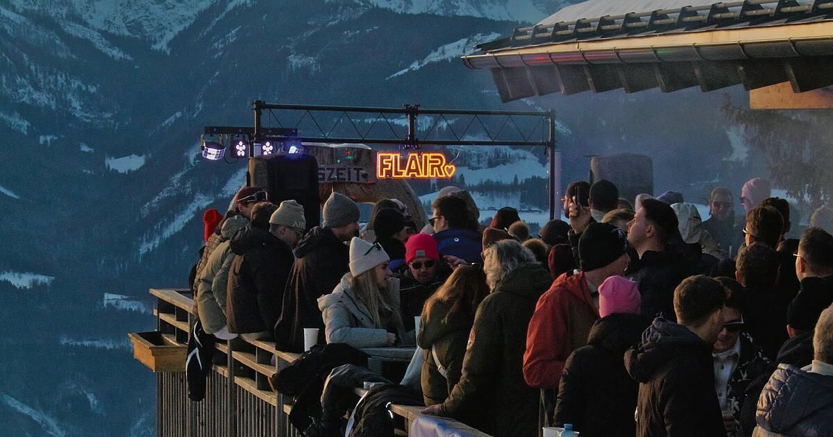 Menschen versammeln sich in einem Außenbereich, wahrscheinlich in einem Skigebiet, unter einem hellen Licht. Der Bereich hat ein Holzgeländer, einen Tisch und Jacken. Sie alle schauen nach oben, möglicherweise auf den Berg.