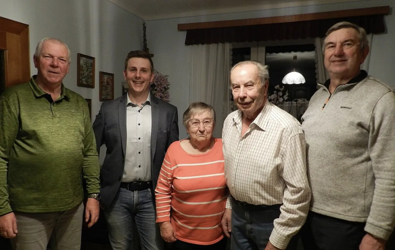 Eine Gruppe von Menschen steht lächelnd zusammen. Ein älterer Mann und eine ältere Frau stehen in der Mitte, mit einer jüngeren Frau und einem jüngeren Mann auf beiden Seiten. Im Hintergrund befindet sich ein Fenster mit Vorhängen und gerahmte Bilder an der Wand.