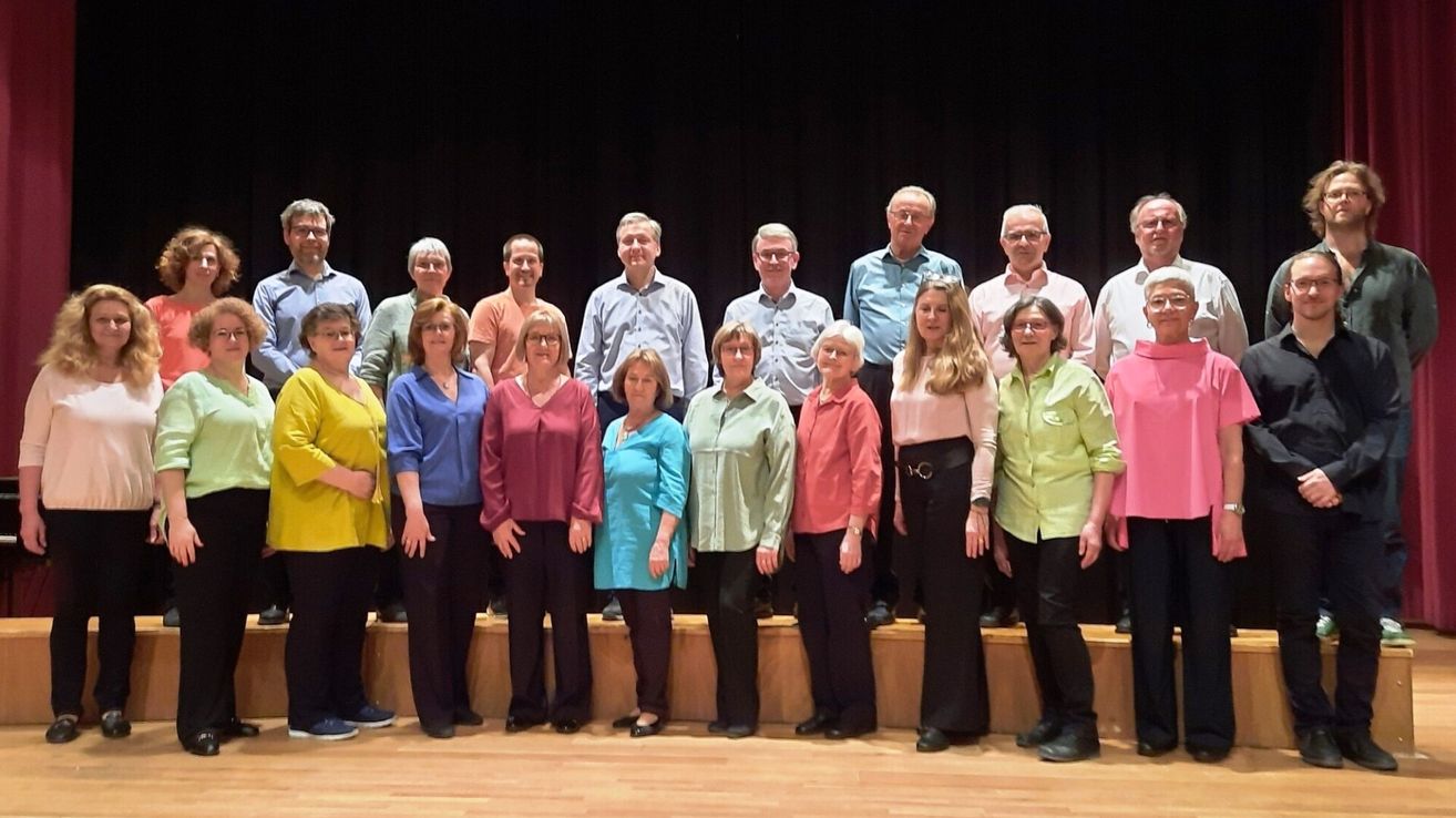 Eine Gruppe von Menschen in passenden Shirts steht auf einer Bühne und lächelt für ein Foto. Die Bühne ist aus Holz, mit einem schwarzen Vorhang im Hintergrund.