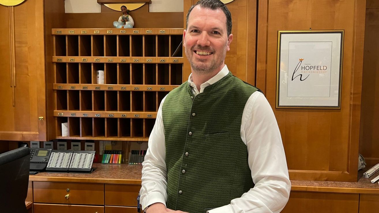 Ein Mann steht lächelnd vor einem hölzernen Empfangstresen und trägt ein weißes Hemd und eine grüne Weste mit Taschen. Hinter ihm befindet sich ein hölzerner Schrank mit beschrifteten Fächern und ein Bilderrahmen an der rechten Wand.