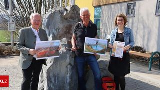 Drei Personen stehen draußen, halten Papiere mit Bildern in der Hand, in der Nähe eines großen Felsens und eines Gebäudes mit Glasfenstern.