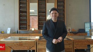 Ein Mann in einem schwarzen Anzug und einer schwarzen Mütze steht in einem Raum mit Holzmöbeln und Regalen voller Brillen.