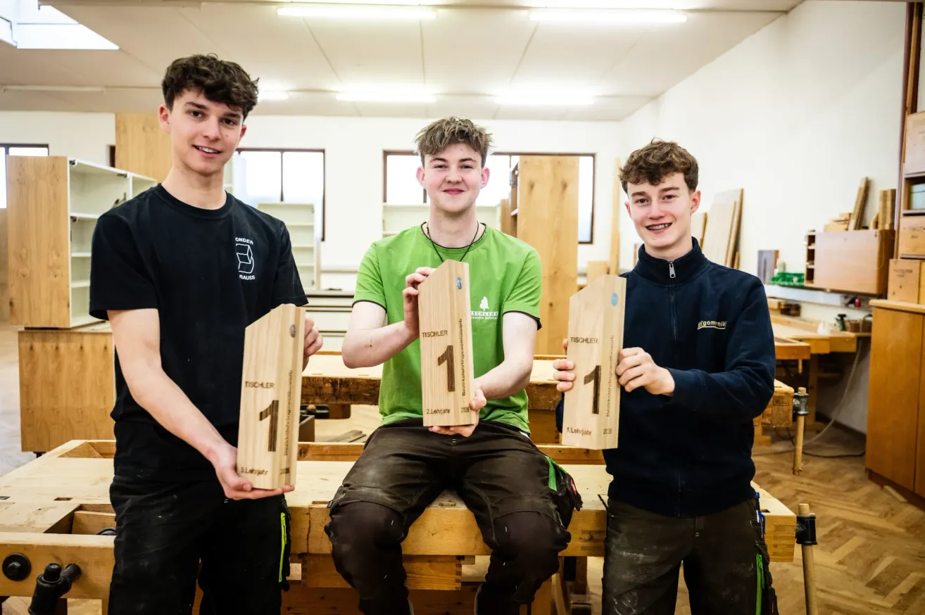 Drei junge Männer stehen zusammen und halten Holztafeln, wahrscheinlich in einer Werkstatt. Sie lächeln und posieren für ein Foto. Jeder hält eine Tafel mit der Nummer 1. Hinter ihnen sind Holzregale und Tische zu sehen.